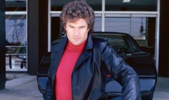A man with curly brown hair wearing a black leather jacket and red turtleneck poses confidently in front of a black sports car outside a building.