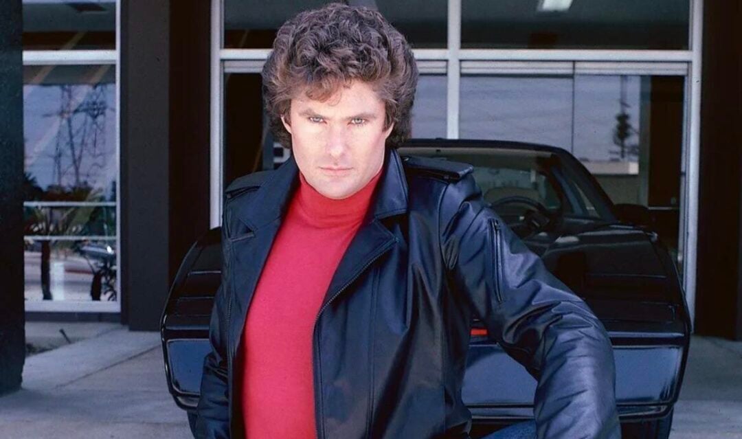 A man with curly brown hair wearing a black leather jacket and red turtleneck poses confidently in front of a black sports car outside a building.