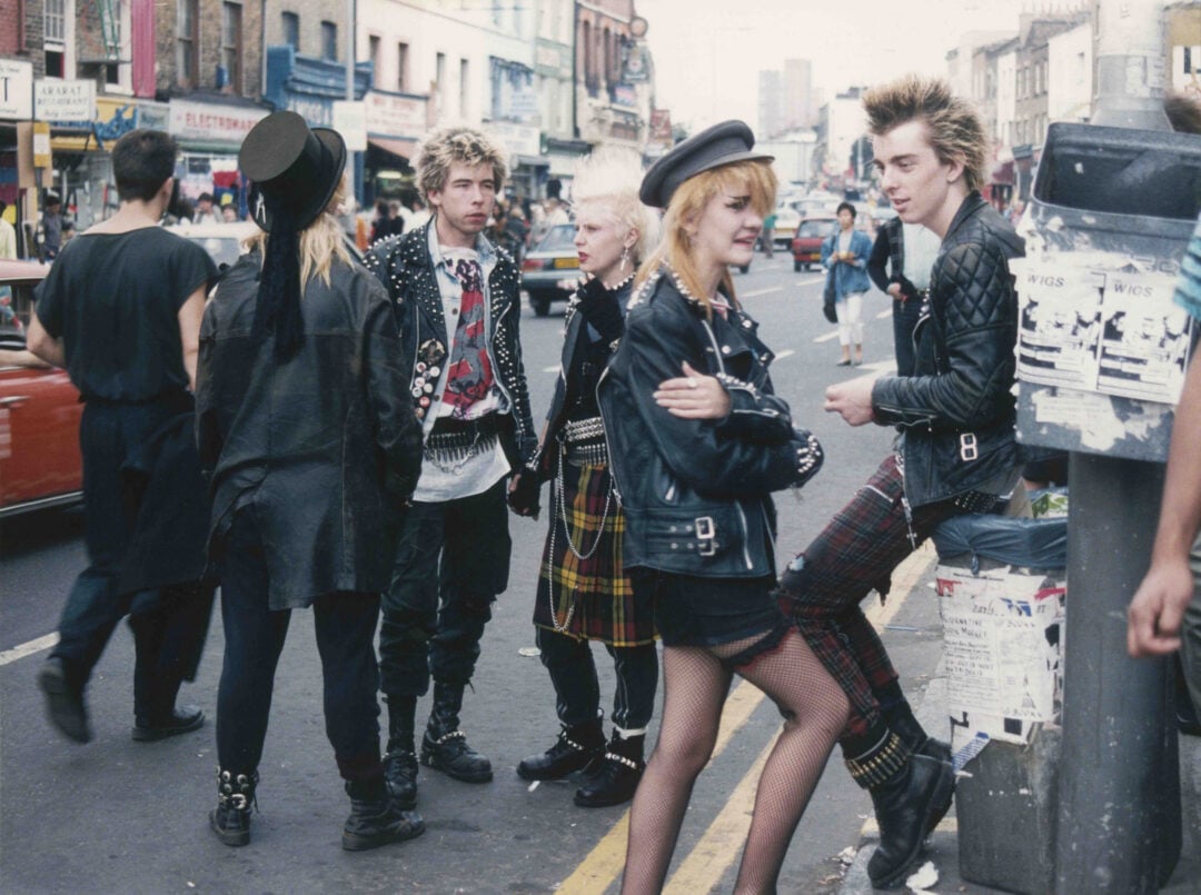 A group of young people dressed in punk fashion—leather jackets, tartan skirts, and spiked hair—stand and chat on a busy city street lined with shops and posters.