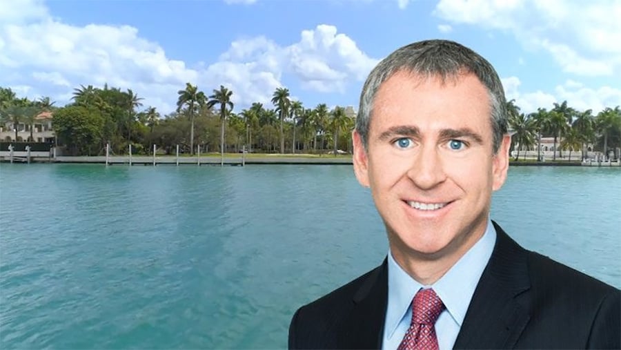 A man in a suit and tie smiles in the foreground with a waterfront, palm trees, and houses visible across the water under a partly cloudy sky.
