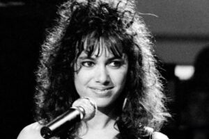 A woman with curly dark hair and bangs stands at a microphone, smiling slightly. She wears a sleeveless, glittery top. The image is in black and white, with a blurred background.