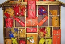 A wooden, house-shaped Advent calendar holds small, wrapped gifts in red, orange, and yellow paper, each tied with gold or silver string and decorated with stars, filling the cubbyholes.
