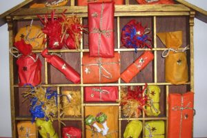 A wooden, house-shaped Advent calendar holds small, wrapped gifts in red, orange, and yellow paper, each tied with gold or silver string and decorated with stars, filling the cubbyholes.