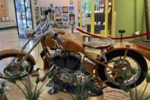 A custom motorcycle with a wooden frame and details is displayed indoors, surrounded by potted plants. A small sign in front provides information about the bike. Artworks and display cases are visible in the background.