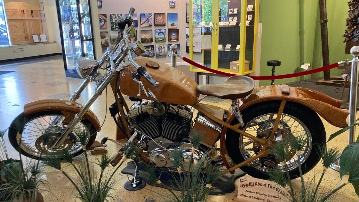 A custom motorcycle with a wooden frame and details is displayed indoors, surrounded by potted plants. A small sign in front provides information about the bike. Artworks and display cases are visible in the background.