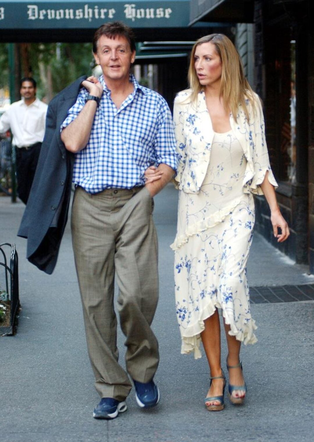 A man in a blue checkered shirt and khaki pants walks arm in arm with a woman in a cream floral dress on a city sidewalk under a green awning that reads "Devonshire House.
