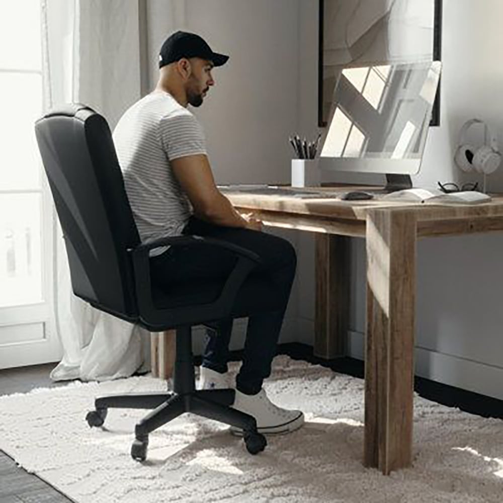 A man wearing a striped shirt, dark pants, and a cap sits on an office chair at a wooden desk, looking at a computer monitor in a bright, modern room with a white rug and large window.