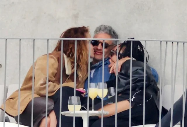 Three people are sitting closely together on a couch outdoors, smiling and relaxing. Two of them are leaning in toward the person in the middle. There are drinks on a small table in front of them. A railing separates them from the camera.