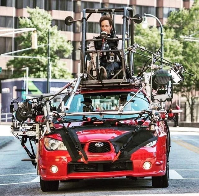 A red car is rigged with cameras, lights, and metal frames, while a person sits in a mounted seat above the car roof, holding steering controls. The setup appears to be for filming or special effects, on a city street.