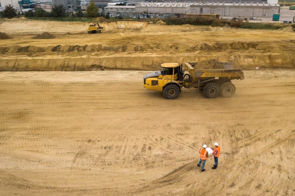 Aerial view of a construction site with a large yellow dump truck and two workers in orange safety vests and helmets standing on sandy ground, with industrial buildings in the background.