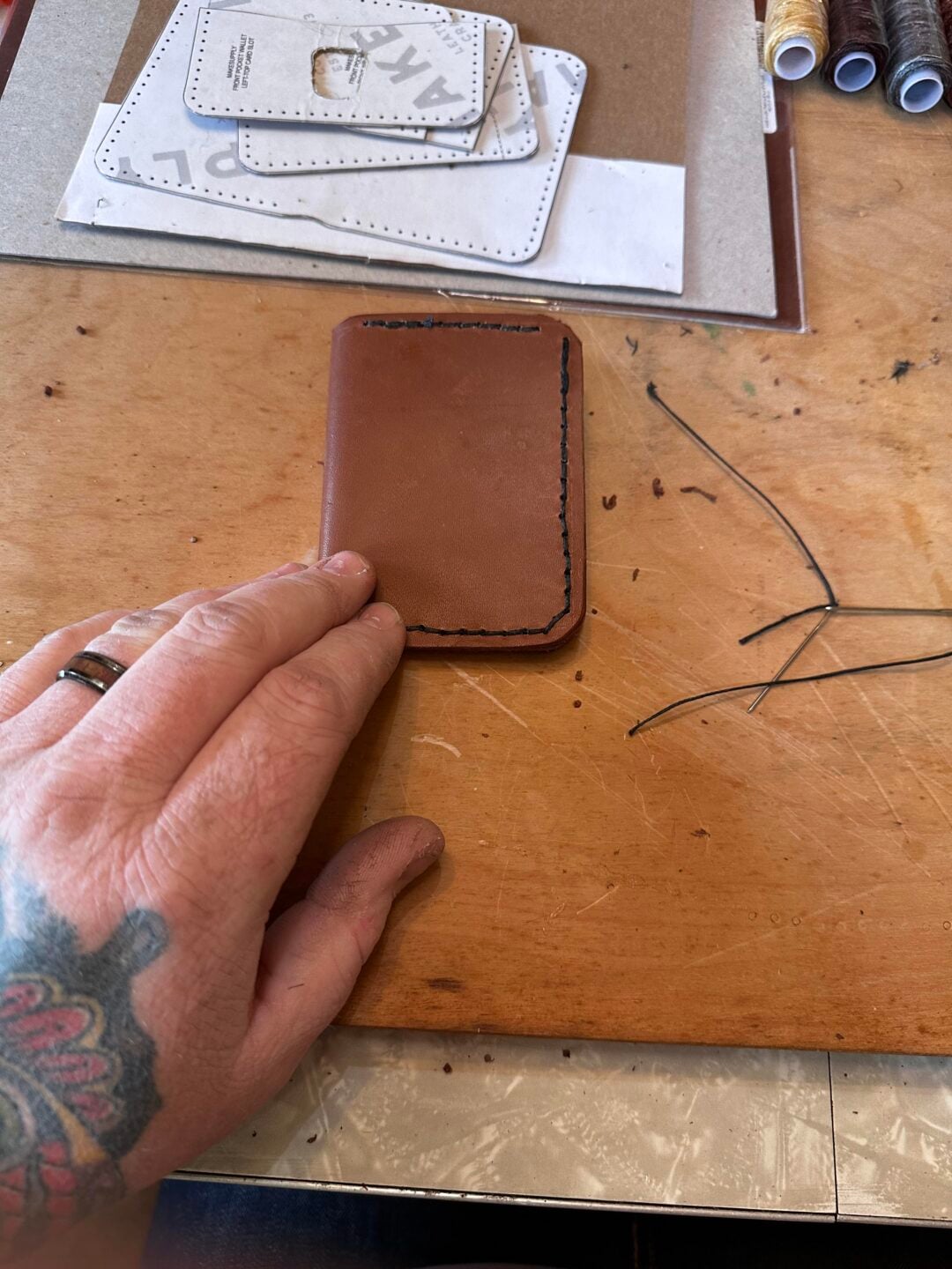 A tattooed hand holds a partially stitched brown leather wallet on a wooden workbench, with black thread, needles, and leather crafting materials nearby.
