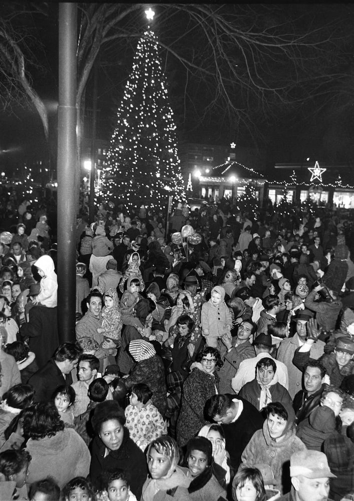 A large crowd of people, including many families and children, gather outdoors at night in front of a tall, decorated Christmas tree with lights. Holiday lights and festive decorations are visible in the background.