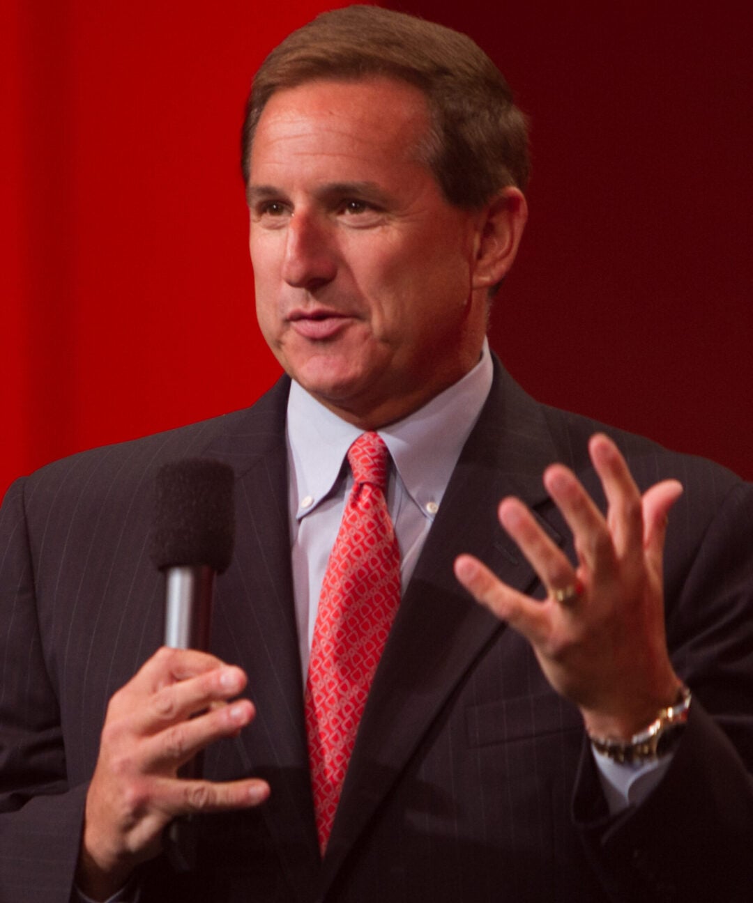 A man in a dark suit, light blue shirt, and red patterned tie speaks into a handheld microphone, gesturing with his other hand, against a red background.