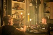 Two boys sit at a kitchen table illuminated by candlelight, eating dinner. Shelves with food and dishes are visible in the background, creating a cozy and intimate atmosphere.