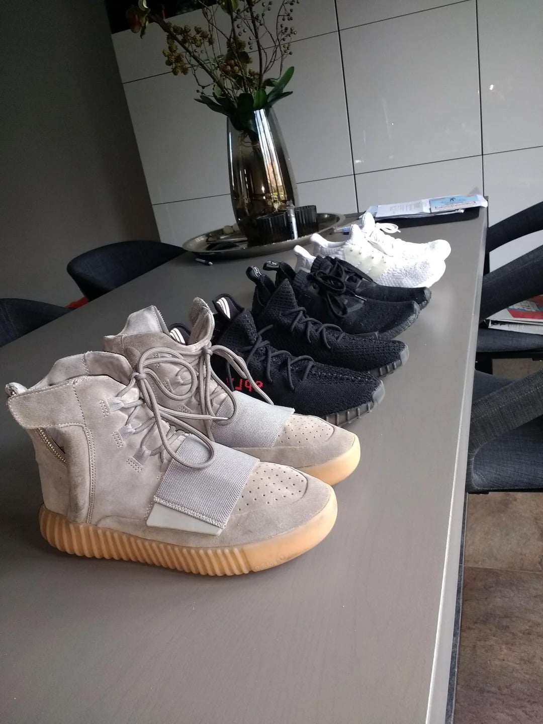 Five pairs of sneakers, including light grey high-tops and several low-top black and white pairs, are neatly lined up on a modern gray table in a dining area with a vase of flowers in the background.
