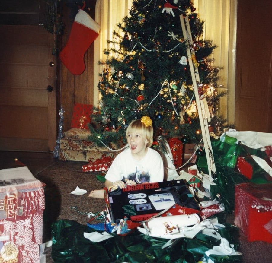 A child with blonde hair and a yellow bow sits on the floor, smiling, surrounded by unwrapped gifts and paper in front of a decorated Christmas tree with lights and ornaments.