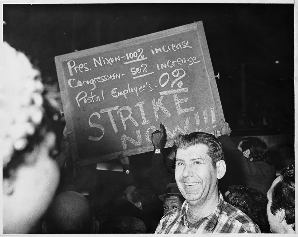 A man stands smiling in a crowd as people hold up a protest sign reading, “Pres. Nixon—100% increase, Congressmen—50% increase, Postal Employee’s—0%. STRIKE NOW!!!”