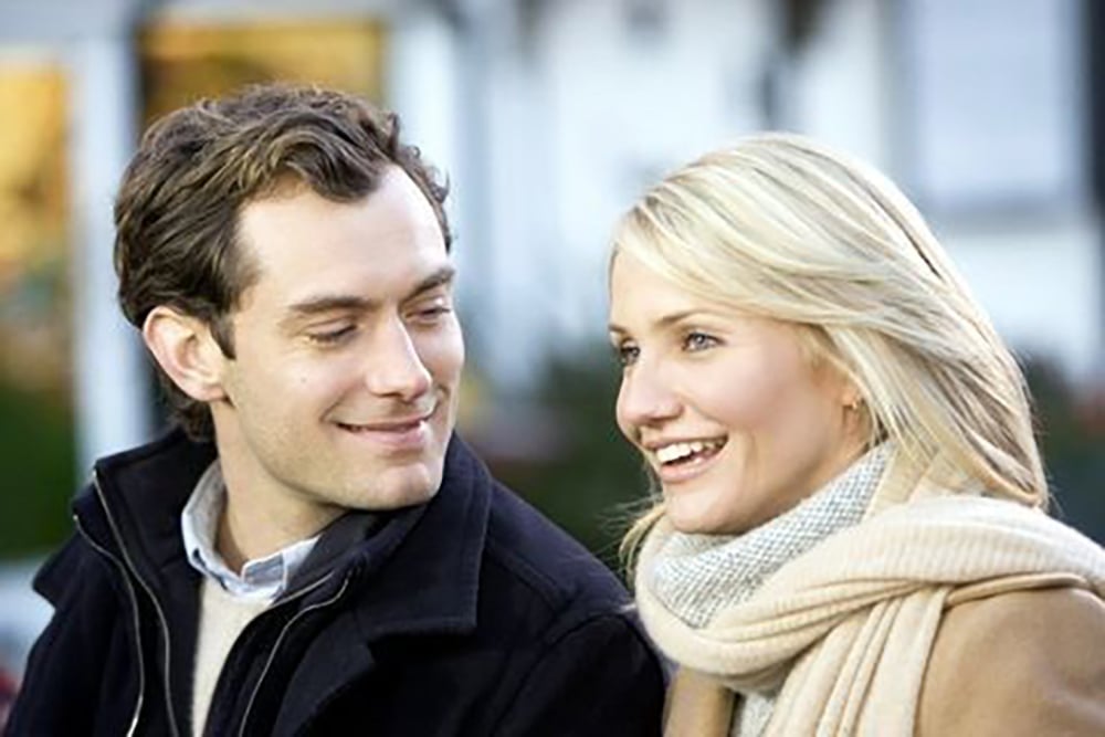 A man and a woman sit close together outdoors, smiling and talking. The woman is wearing a beige scarf and coat, while the man is wearing a dark coat and looking at her with a warm expression.