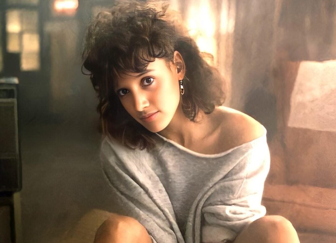 A woman with curly hair in an off-the-shoulder sweater sits on a cushion, legs apart, wearing red heels. The name "Jennifer Beals" appears at the bottom in white script.