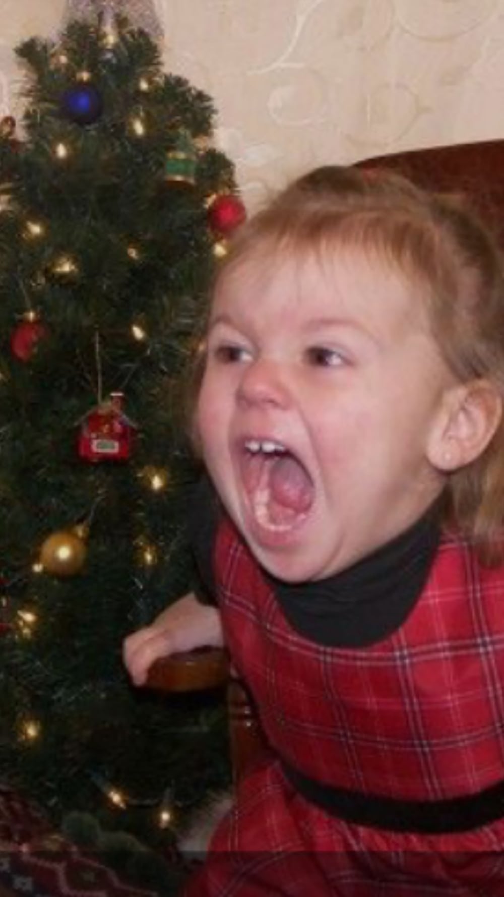 A young girl in a red plaid dress sits beside a decorated Christmas tree, with her mouth wide open in an expressive, playful scream or yell.