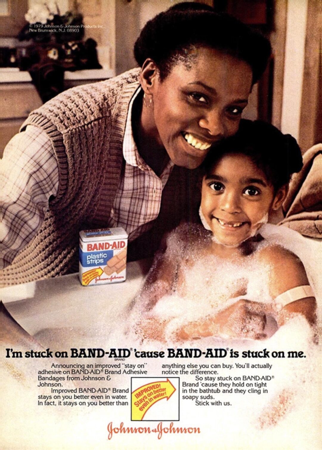 A smiling woman hugs a young girl taking a bubble bath. The girl has a BAND-AID on her arm. A box of BAND-AID bandages sits nearby. Text promotes BAND-AID’s stickiness. Johnson & Johnson branding appears at the bottom.