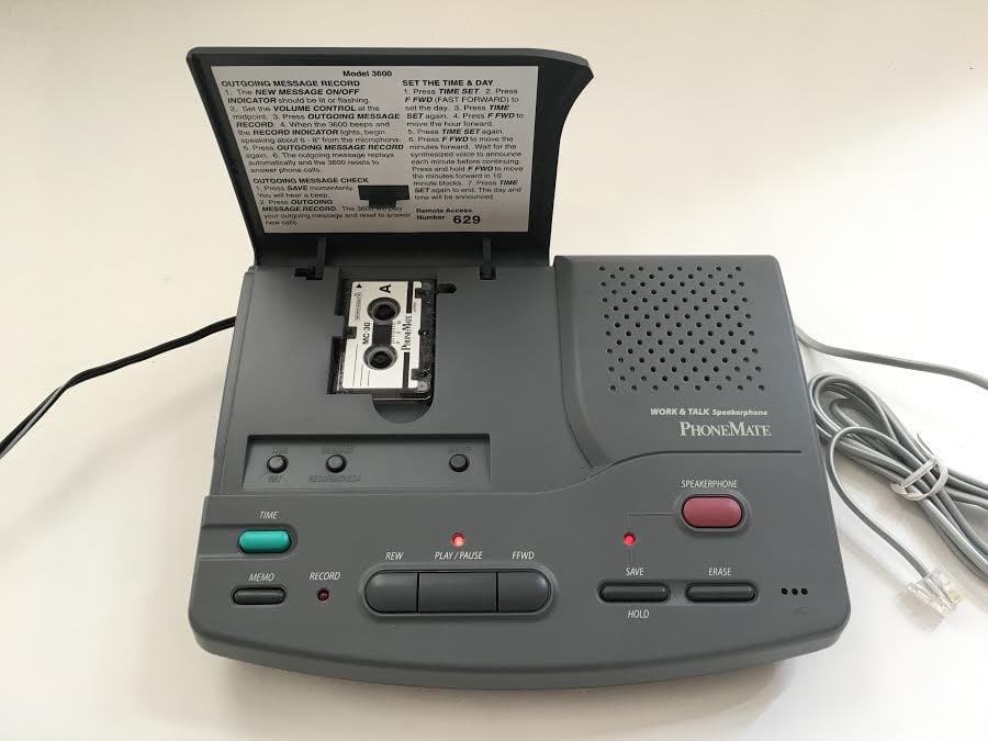 A vintage PhoneMate answering machine with an open cassette compartment, visible tape inside, various buttons for functions like play, record, forward, and a coiled phone cord attached on the right side.