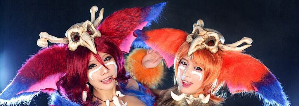 Two people in vibrant, furry costumes with large ears, face paint, and bone headpieces pose cheerfully against a dark background, imitating fantasy or game characters. Bright colors like red, orange, and blue dominate their outfits.
