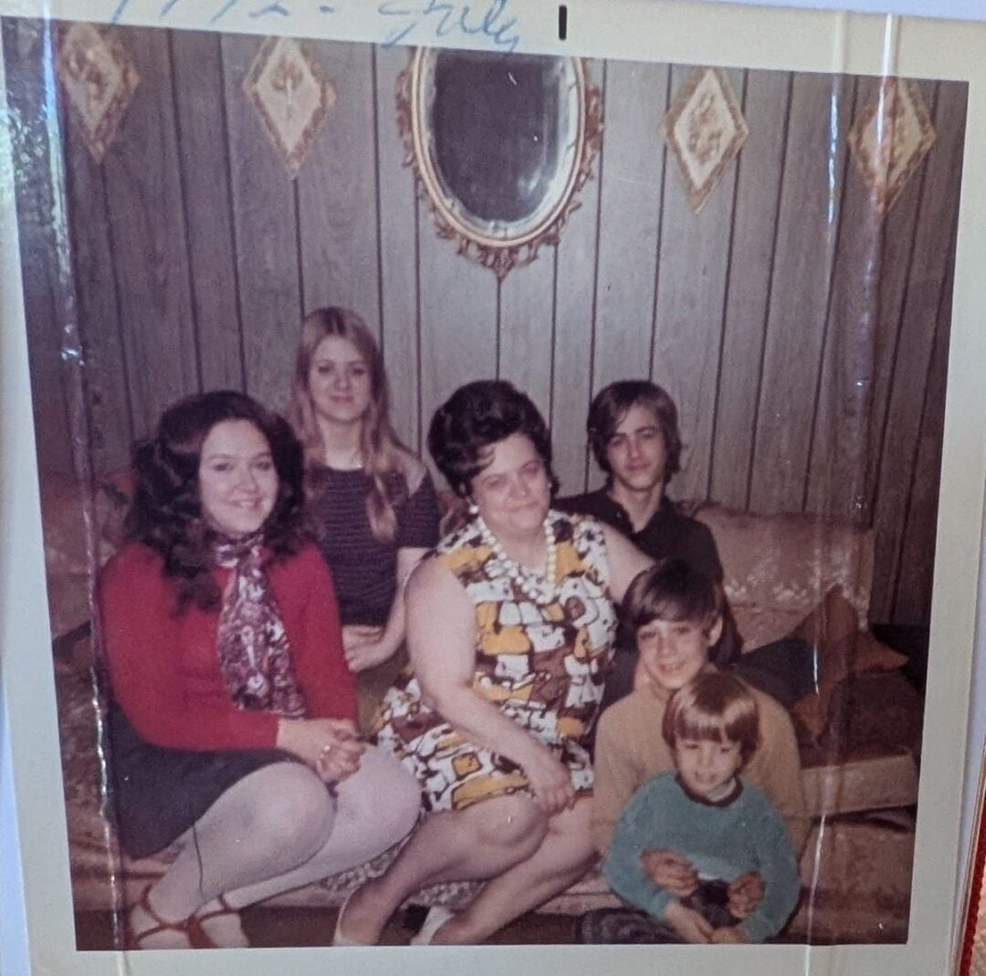 A vintage photo of six people sitting on a couch in a wood-paneled living room, including four children and two women. A small oval mirror hangs on the wall above them. All are looking at the camera.