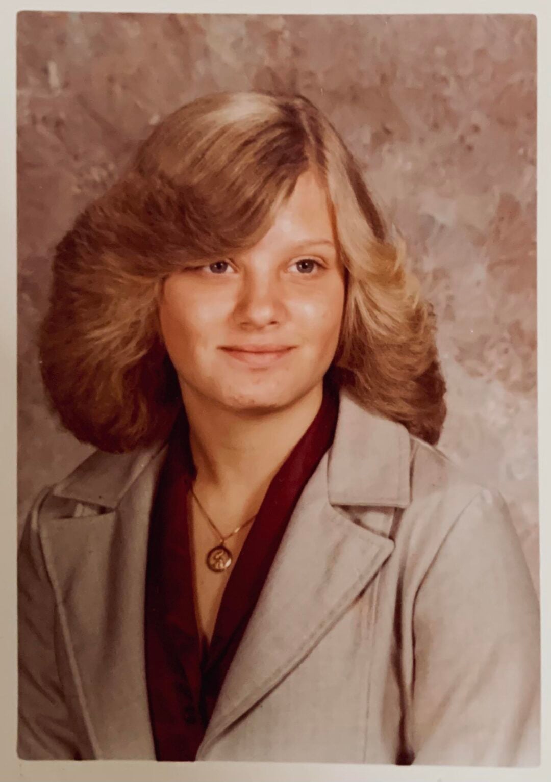 A young woman with feathered, shoulder-length blonde hair wears a light gray blazer over a burgundy blouse and a necklace with a circular pendant, posing for a formal studio portrait against a mottled background.