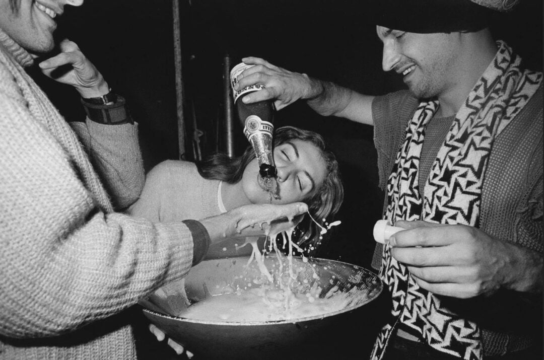 Three people celebrate together. One person pours a bottle of champagne into a large bowl while a woman leans in to drink from the bubbly foam, smiling. Another person holds the bowl steady. The mood is joyful and festive.