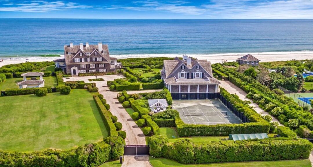 Aerial view of two large beachfront mansions with manicured lawns, a tennis court, gazebo, and direct access to a sandy beach and the ocean under a blue sky.