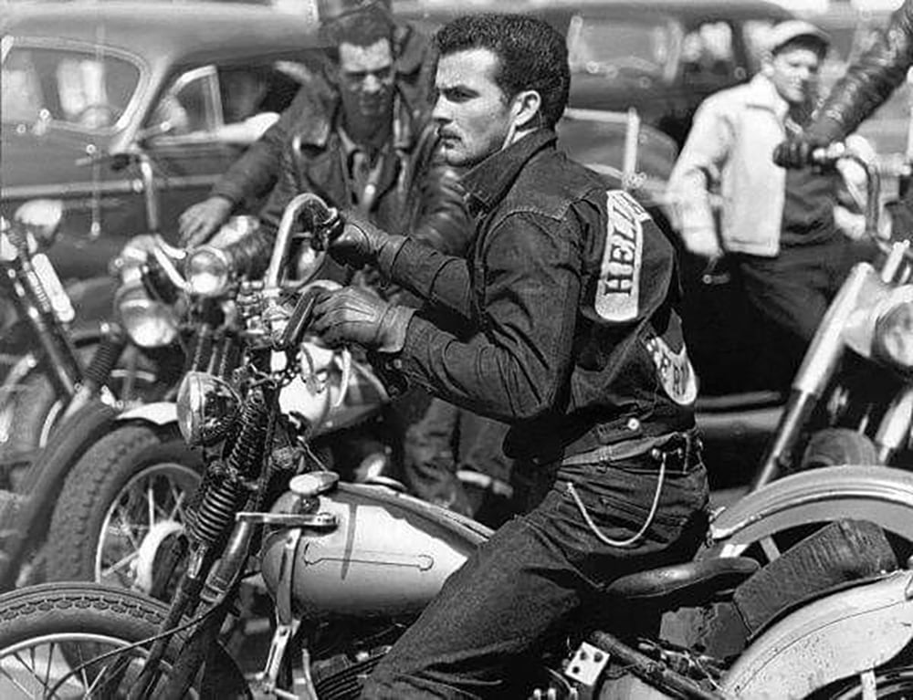 A man in a denim jacket sits on a vintage motorcycle among a group of bikers and parked cars; people in the background are also gathered around motorcycles. The scene appears to be from the mid-20th century.