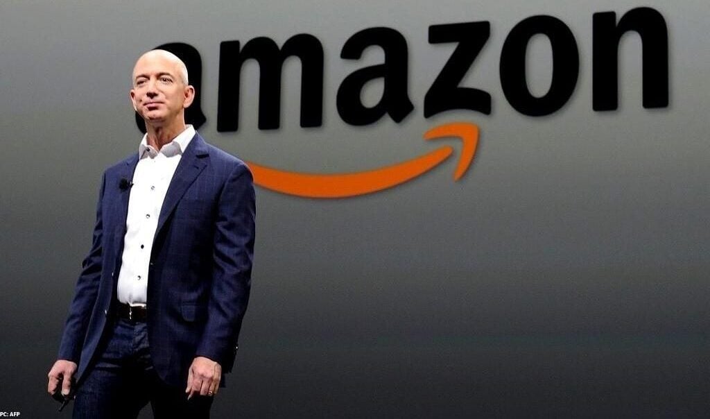 A man in a dark suit and white shirt stands on stage in front of a large Amazon logo projected on a light background.