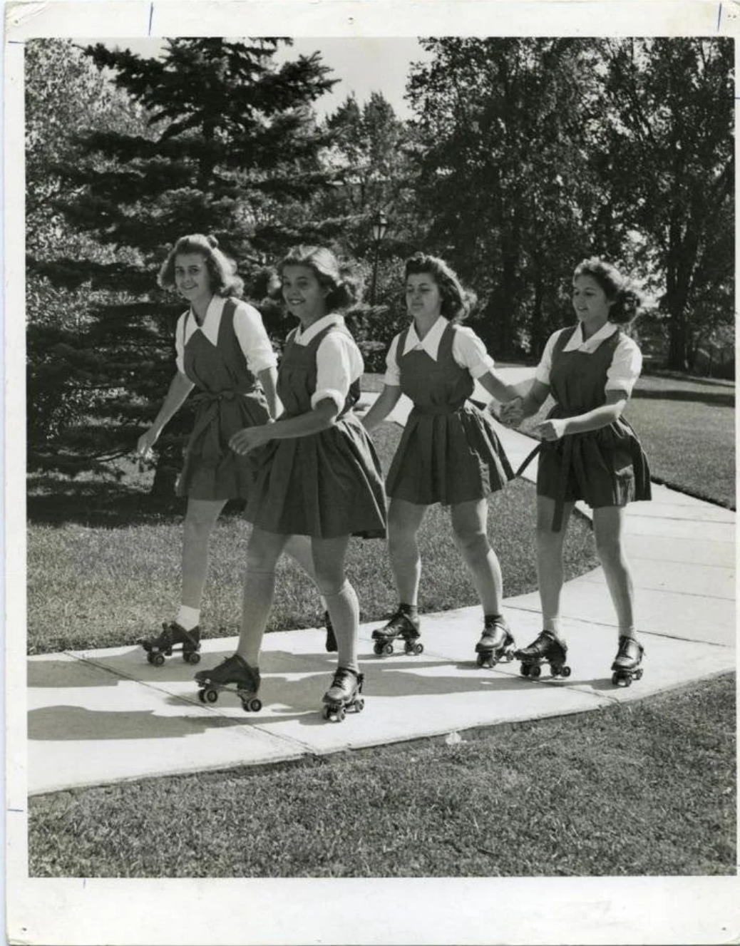 Four young women wearing matching uniforms roller skate together on a sidewalk in a park-like setting, holding hands or arms, with trees and grass in the background. The image appears black and white.