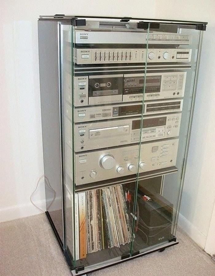 A glass cabinet holds a vintage Sony stereo system with multiple stacked components and a collection of vinyl records stored on the bottom shelf. The unit is placed against a white wall on a carpeted floor.