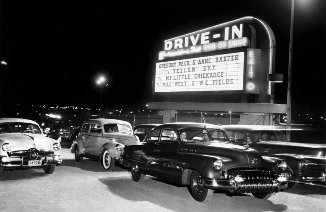 Black-and-white photo of a drive-in theater at night, with vintage cars parked in front of a lit marquee listing several movies, including “Yellow Sky” and “My Little Chickadee,” starring Gregory Peck and Anne Baxter.