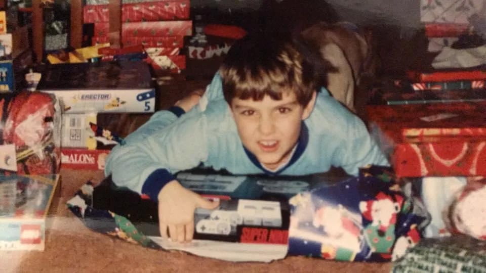 A young boy in pajamas excitedly hugs a newly unwrapped Super Nintendo console on Christmas morning, surrounded by festive wrapping paper and other presents.