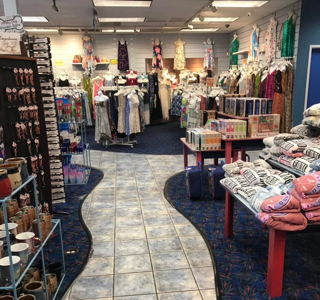 A store interior with dresses on racks, shelves of mugs, souvenirs, and folded towels. A winding tile path runs through the store, and the carpet has a colorful pattern. A sign overhead reads "JUNIORS.