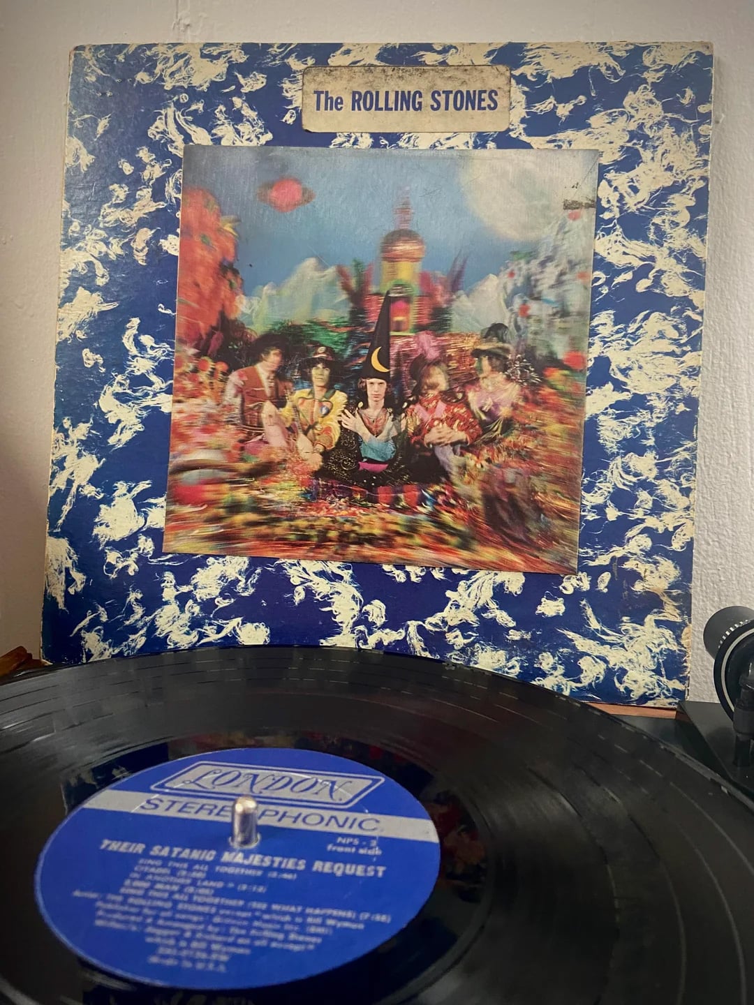 A vinyl record plays on a turntable in front of the colorful, psychedelic album cover of "Their Satanic Majesties Request" by The Rolling Stones. The cover features the band in vibrant, fantastical costumes.