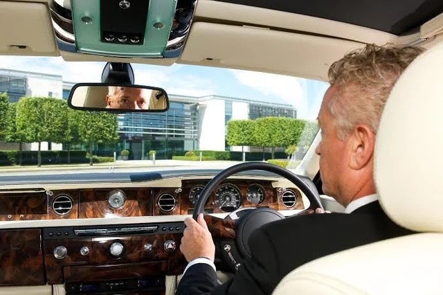 A man with gray hair in a suit drives a luxury car with a wood-finish dashboard, while his reflection is visible in the rearview mirror. Modern buildings and trimmed hedges are seen outside through the windshield.