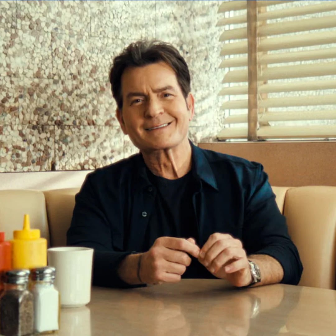 A man with dark hair sits at a diner booth, smiling. He wears a dark shirt and jacket. On the table are condiments, salt, pepper shakers, and a white coffee mug. Blinds and a textured wall are in the background.