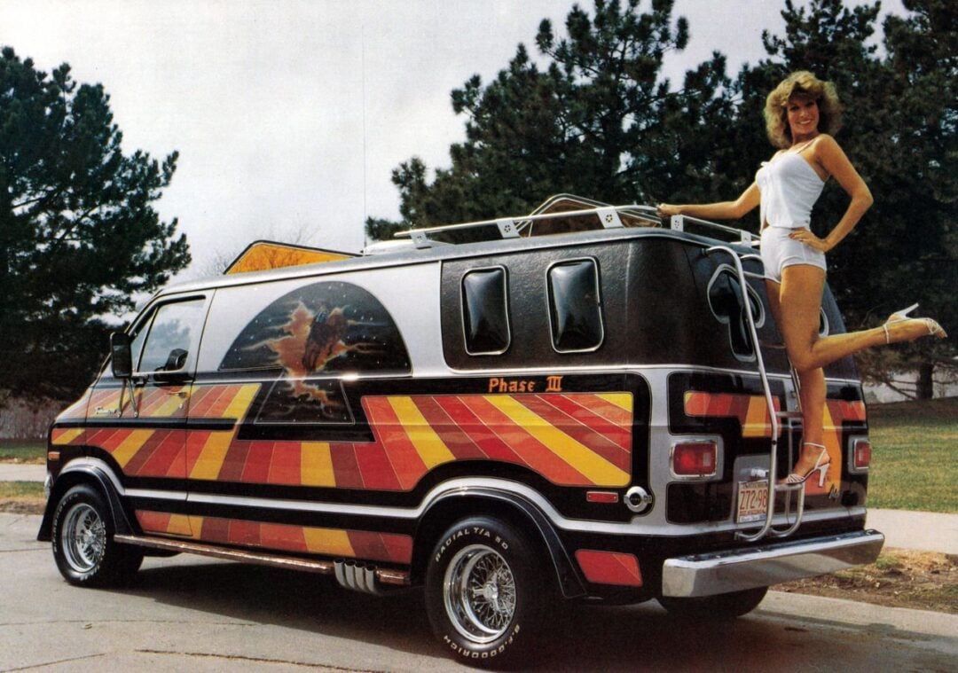 A woman in a white outfit and high heels poses on the rear bumper of a custom black van with colorful red, yellow, and orange stripes and a mural of a lion on the side, parked outdoors near trees.