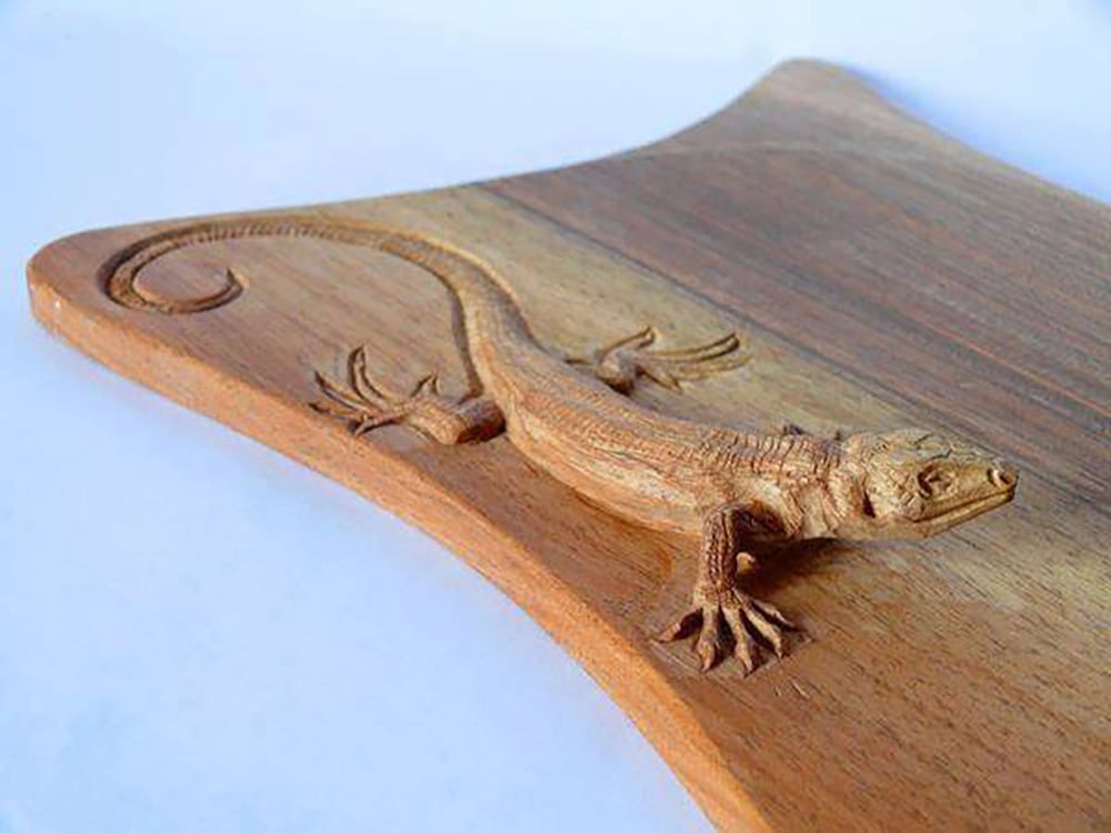 A wooden cutting board with a detailed, three-dimensional carving of a lizard appearing to crawl across its surface. The lizard and board are both made from the same wood.