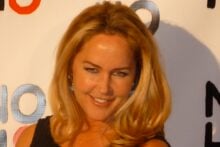 A woman with long blonde hair and blue earrings smiles at an event, standing in front of a white backdrop with the "NOH8" campaign logo in black, red, and gray letters.