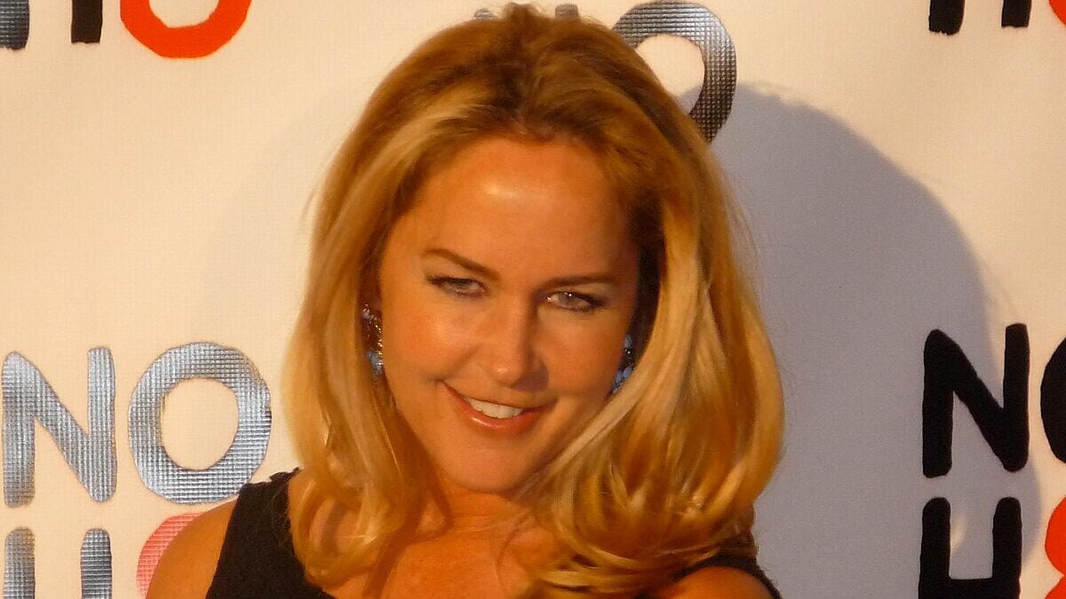 A woman with long blonde hair and blue earrings smiles at an event, standing in front of a white backdrop with the "NOH8" campaign logo in black, red, and gray letters.