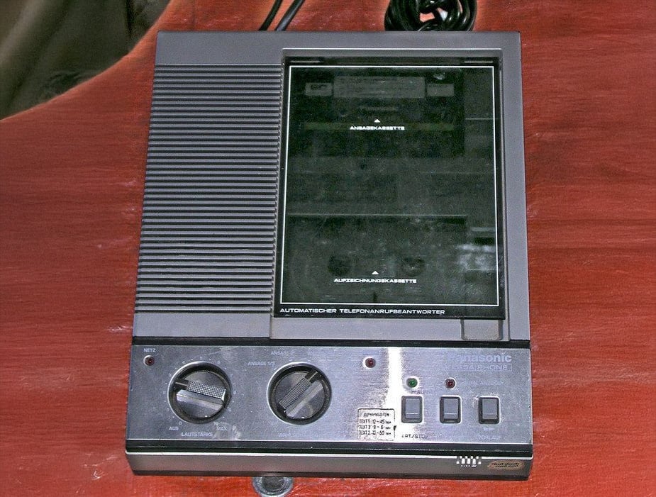 A vintage Panasonic telephone answering machine with a cassette tape deck, two control knobs, buttons, and indicator lights, sitting on a reddish-brown wooden surface.