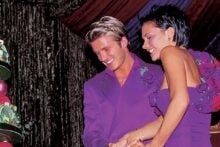 A smiling couple in matching purple outfits cut a large, colorful wedding cake with greenery and red apples on it. They are under a decorated tent, with lush drapery in the background.