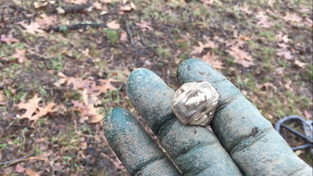 A gloved, dirt-covered hand holds a tarnished, angular ring outdoors; scattered brown leaves and grass are visible on the ground in the background.