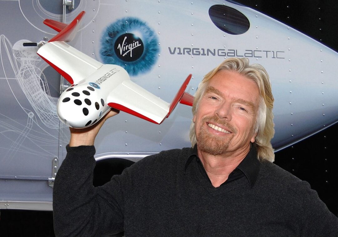 A smiling man with long blond hair holds a model spaceship in front of a Virgin Galactic spacecraft display featuring the company&rsquo;s logo and branding.