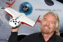 A smiling man with long blond hair holds a model spaceship in front of a Virgin Galactic spacecraft display featuring the company&rsquo;s logo and branding.